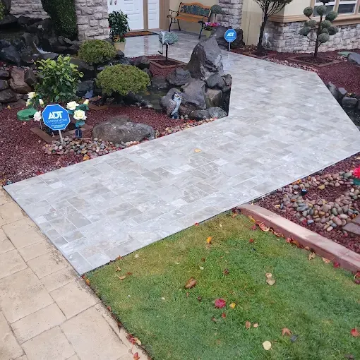 Curved light stone walkway through landscaped front yard with rocks, shrubs, and grass beside a house