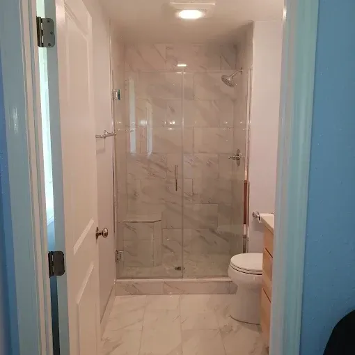 Small bathroom with a glass-enclosed shower, toilet, and beige tile floor, viewed through an open door