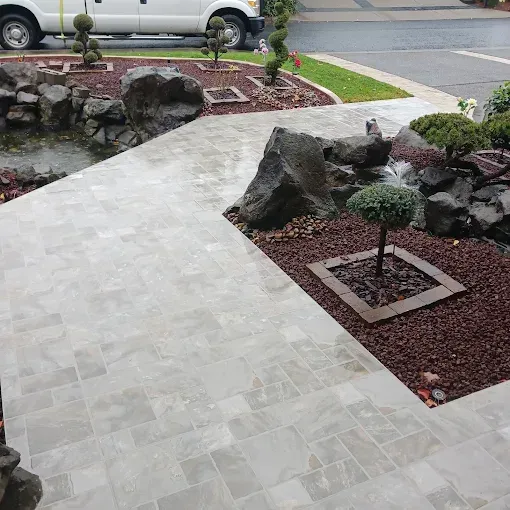 Stone walkway through a landscaped garden with rocks, trimmed shrubs, and a parked van in the background