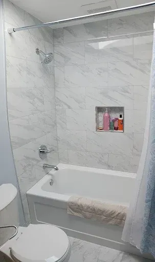 Small white bathroom with a tub-shower, marble-look tiles, toilet, and a shower curtain.