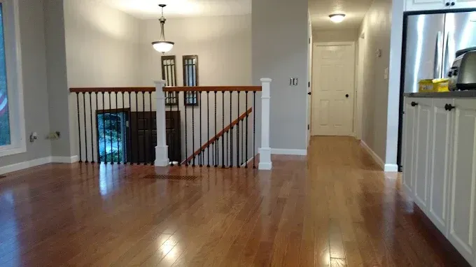 Empty modern living room with hardwood floors, white walls, staircase railing, and kitchen area visible on the right