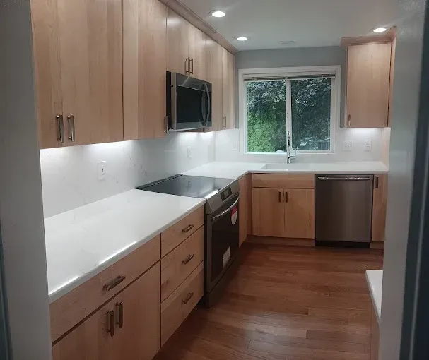 Modern kitchen with light wood cabinets, white counters, stainless appliances, and a window above the sink