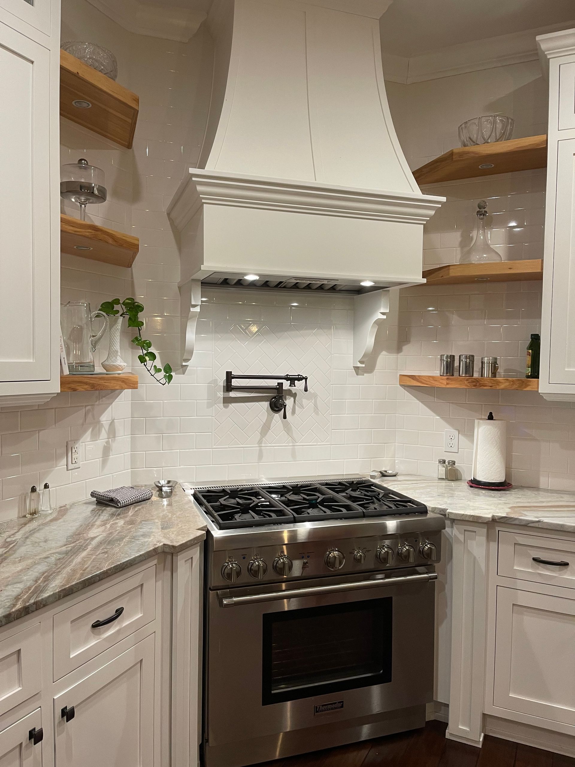 White kitchen with a stainless steel stove, white range hood, and wooden shelves.