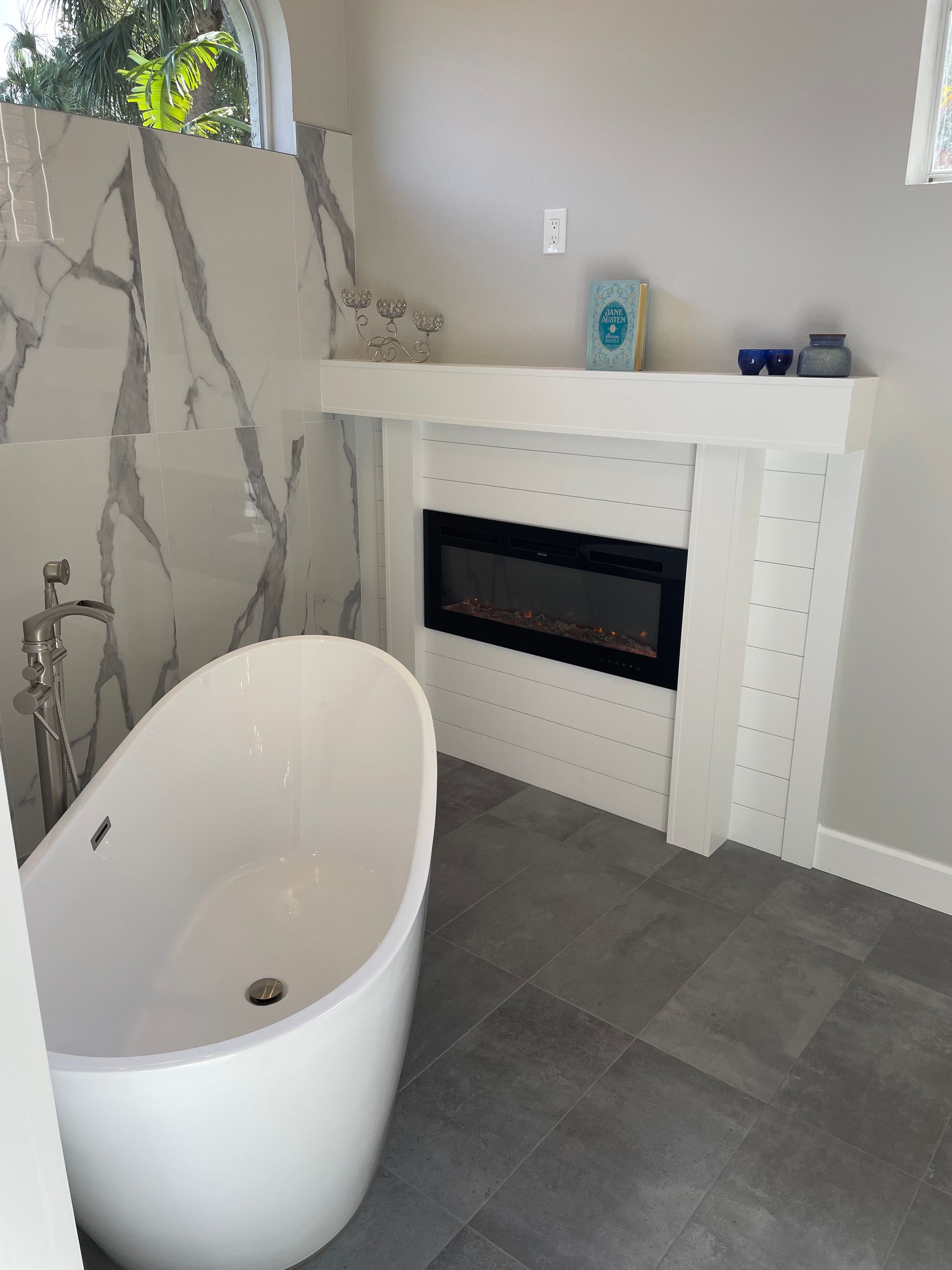 White soaking tub, fireplace with black trim, marble accent wall in a modern bathroom.