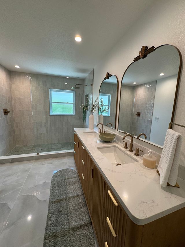 Modern bathroom with dual sinks, gold-framed mirrors, and a large shower. Wooden vanity and grey tiled walls.