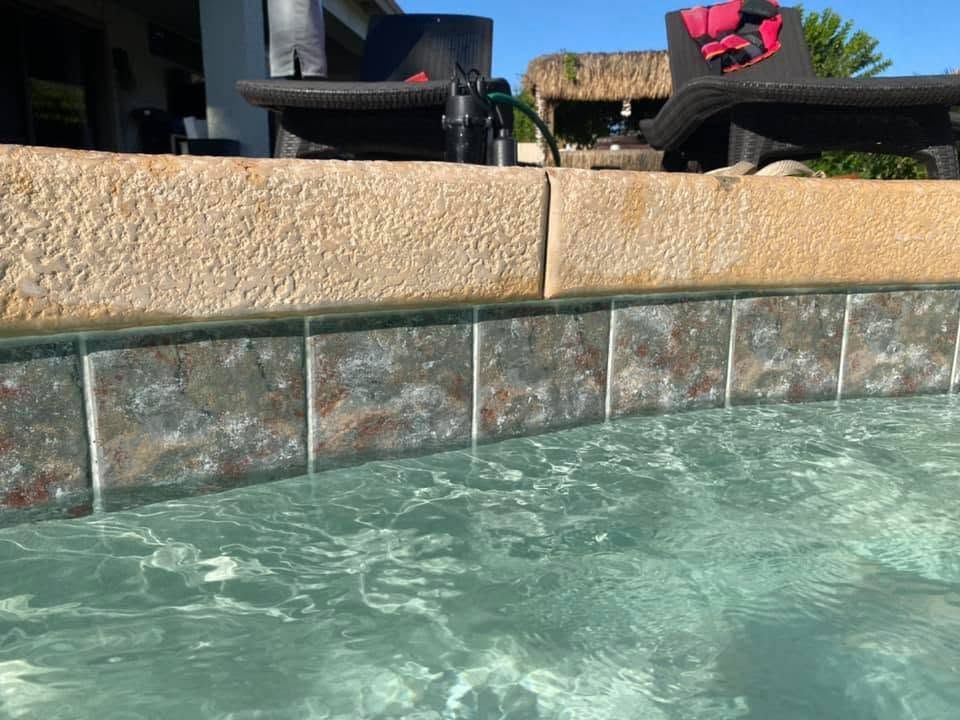 Swimming pool water beside a beige stone ledge with patio chairs in the background