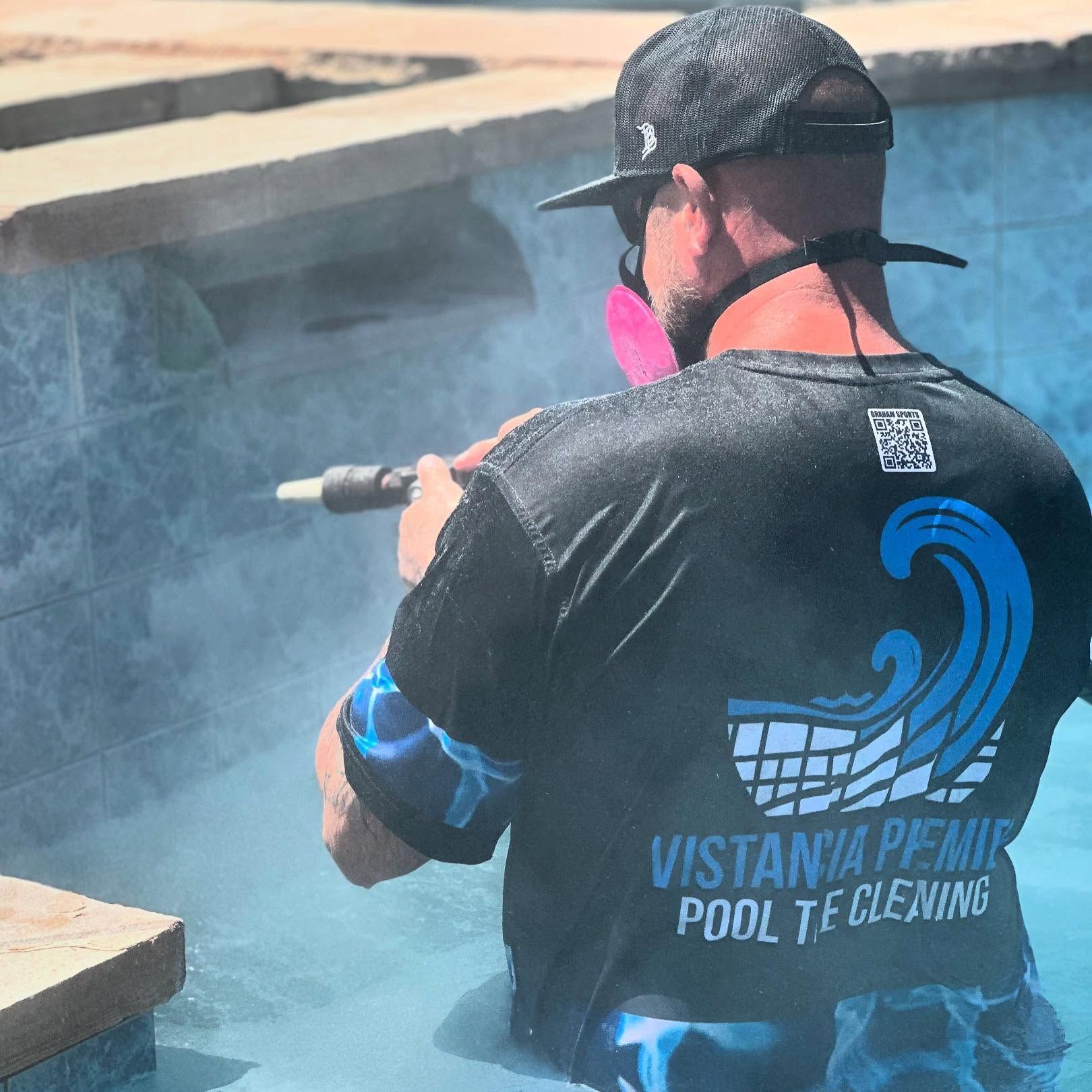 Worker pressure-washing a pool edge with a spray wand, wearing a black shirt and cap, near blue-tinted water.