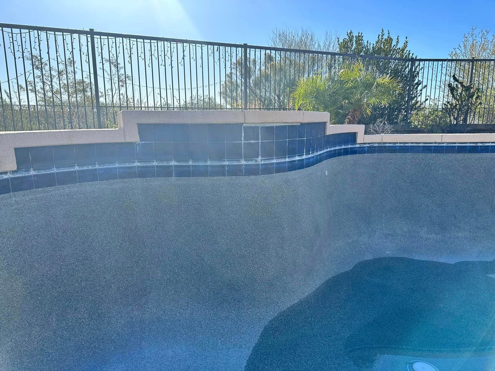 Empty swimming pool with blue-tiled walls, steps, and a fence under bright sunlight
