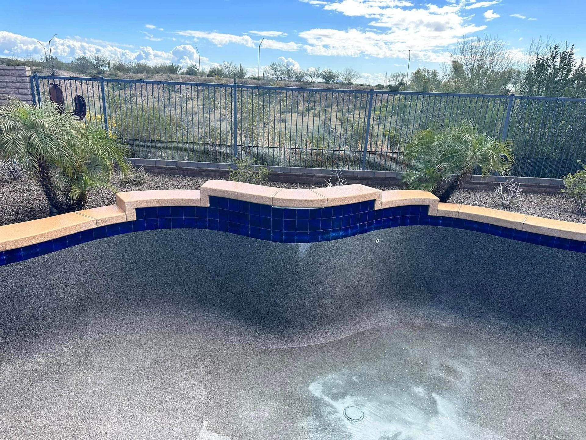 Empty blue pool with coping stones, viewed against a desert landscape and metal fence under a cloudy sky