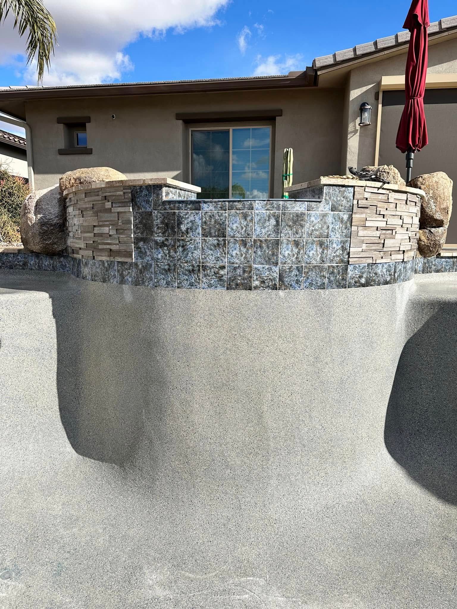 Backyard pool with stone waterfall feature in front of a stucco house and red patio umbrella