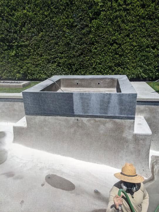 Stone outdoor sink and bench in front of a green hedge, with a straw-hatted person seated at bottom right