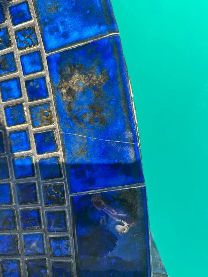 Blue mosaic tile edge beside turquoise water, with small square tiles and weathered grout.