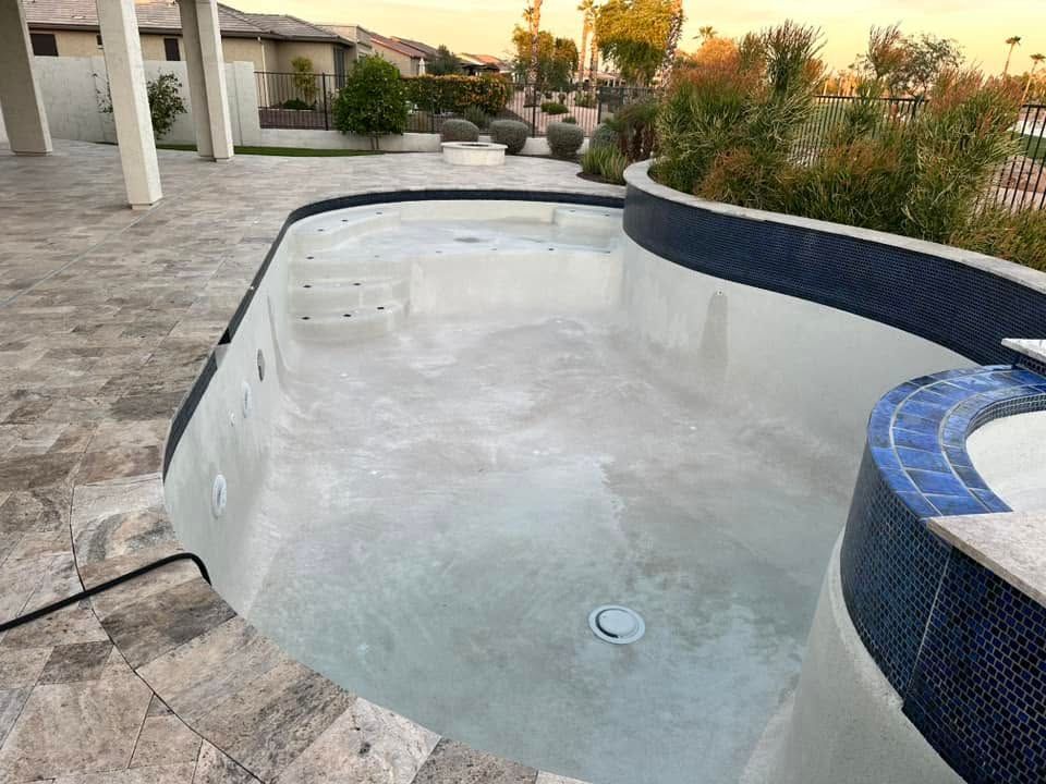 Empty backyard swimming pool with tiled coping and water at the bottom, beside a patio and plants