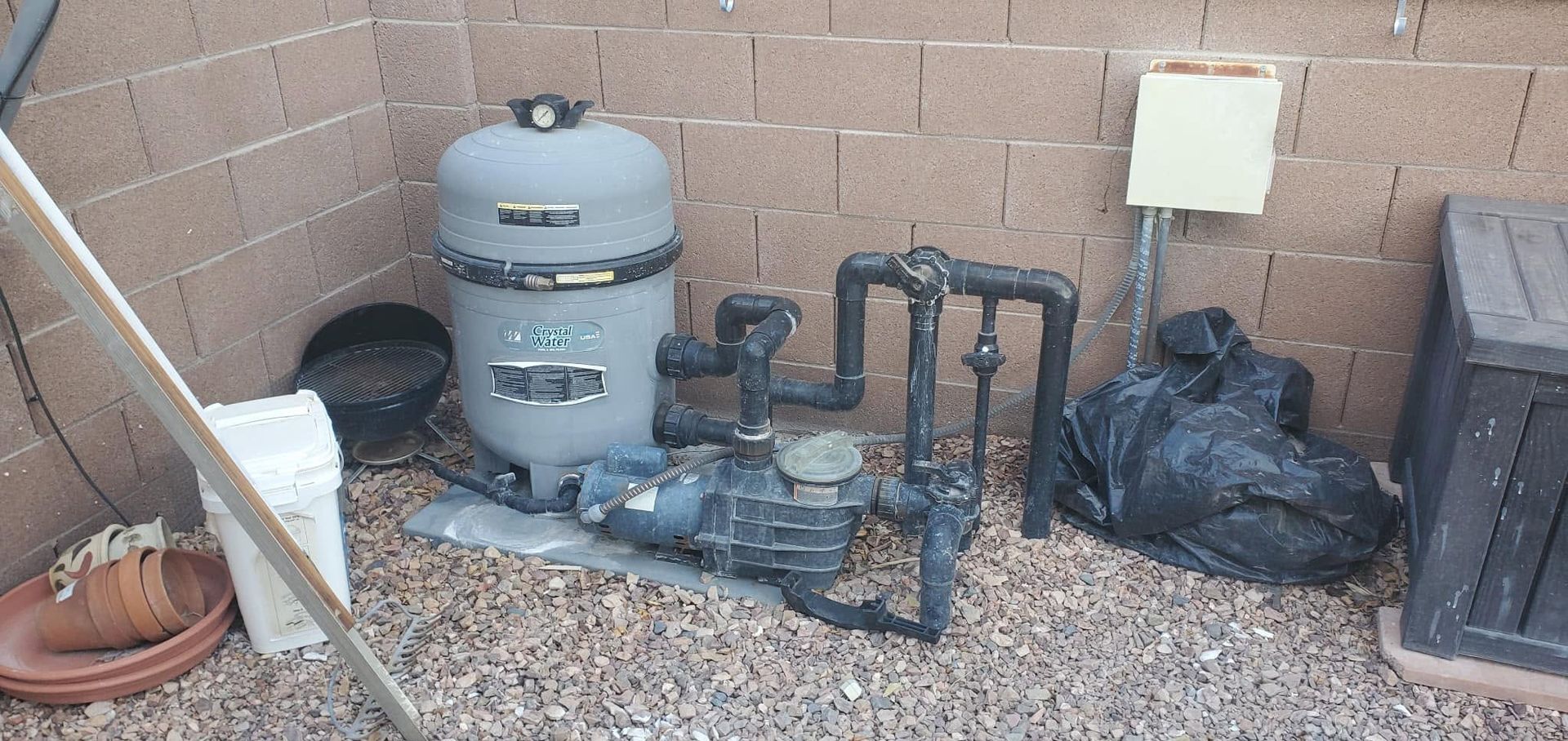 Backyard pool equipment and plumbing against a brick wall, with gravel and scattered bags