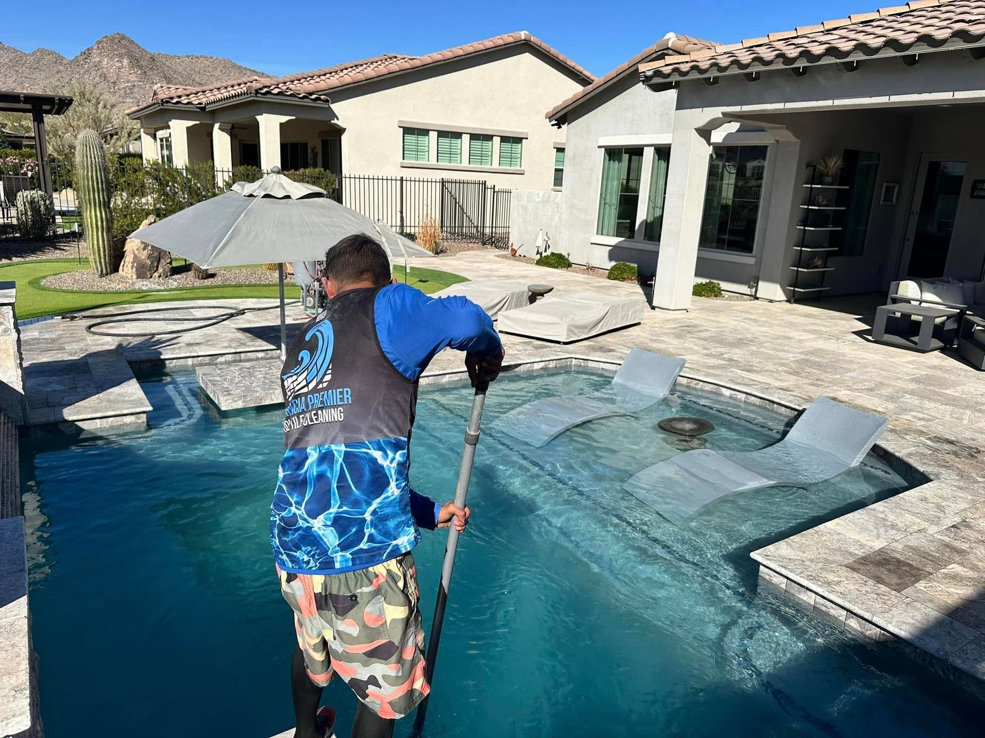 Person cleaning a backyard pool beside a modern home on a sunny day
