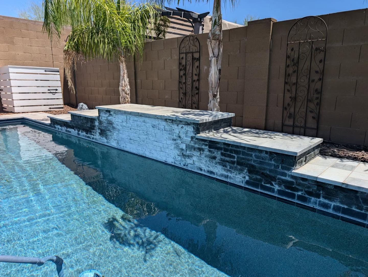 Backyard pool with blue water and a stone waterfall against a tan wall and patio seating