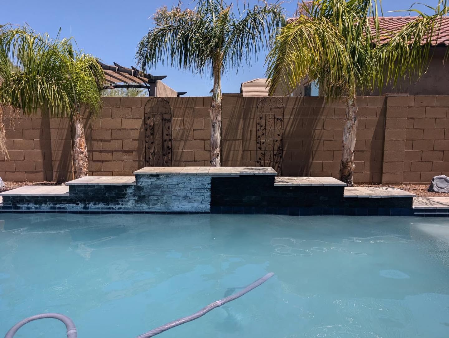 Backyard pool with palm trees, brick wall, and a small raised spa waterfall in bright sunlight