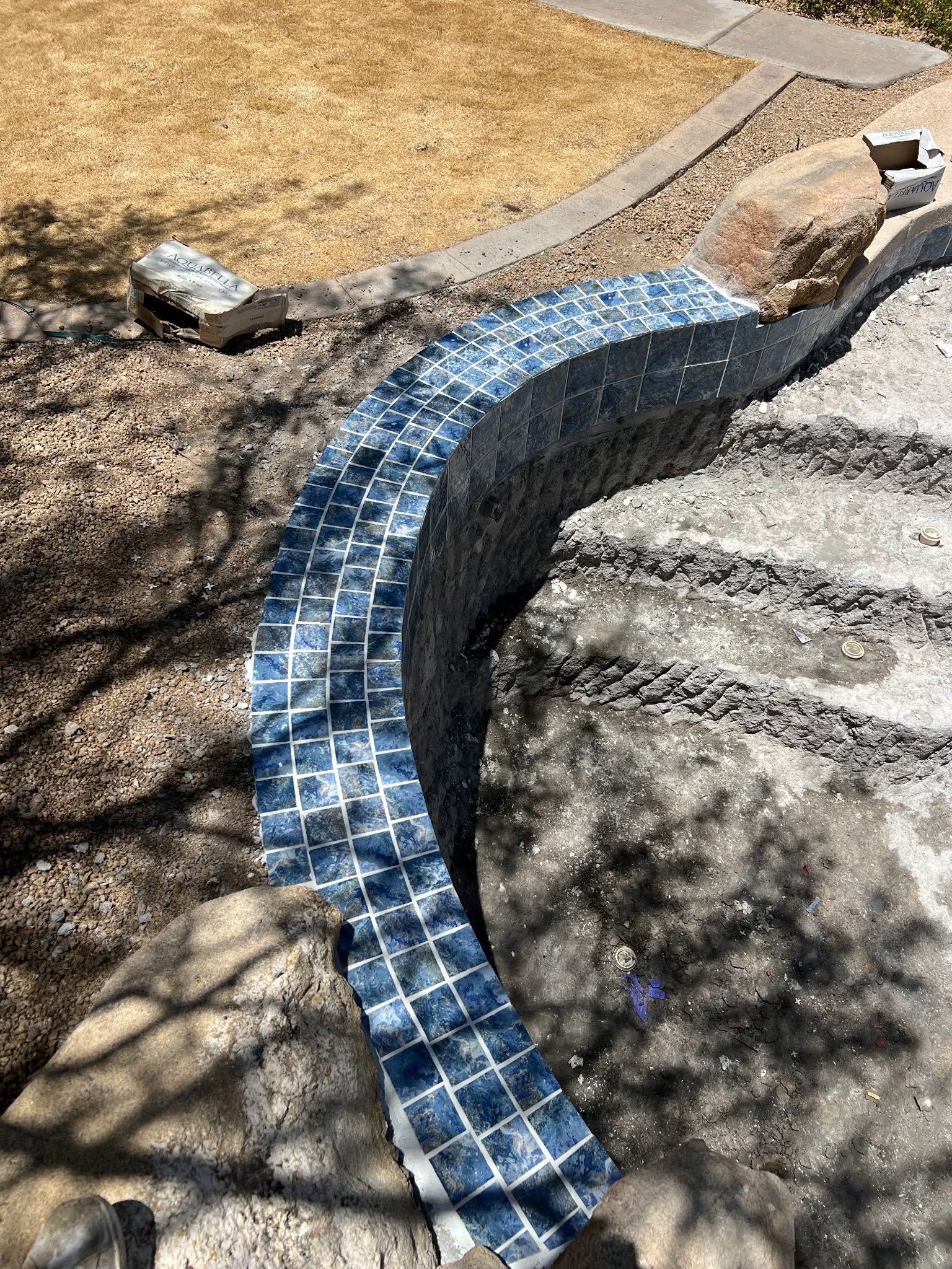 Curved blue-tiled garden border beside a stone path and dry grass, with tree shadows overhead