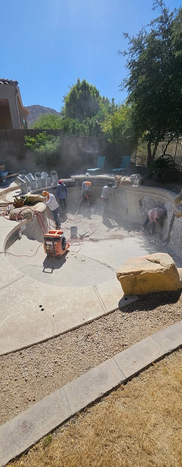 Steaming outdoor hot spring pool with people relaxing in bright sunlight, surrounded by rocks and trees.