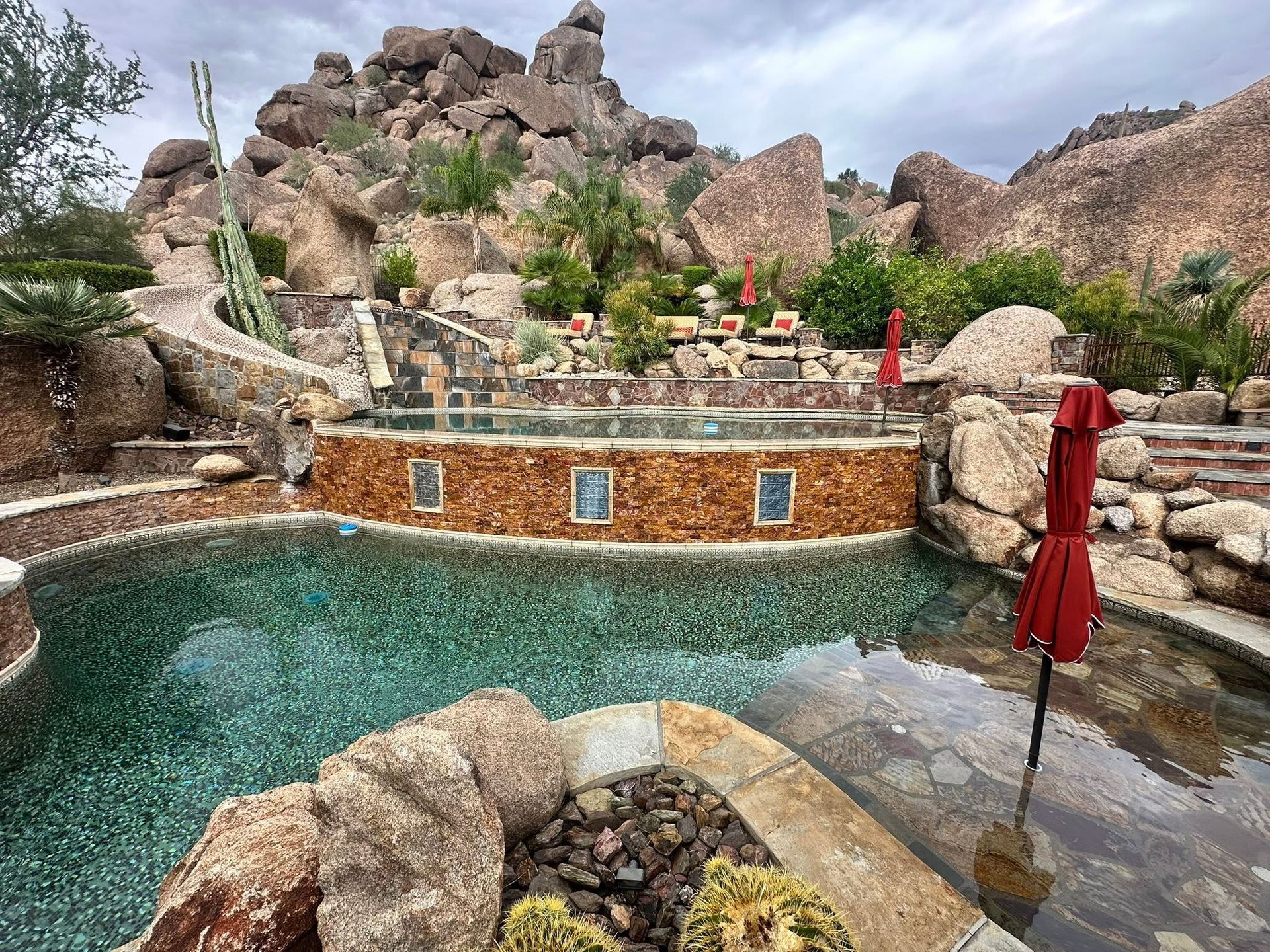 Rocky resort pool with turquoise water, stone hot tub, and red umbrellas beneath a cloudy sky