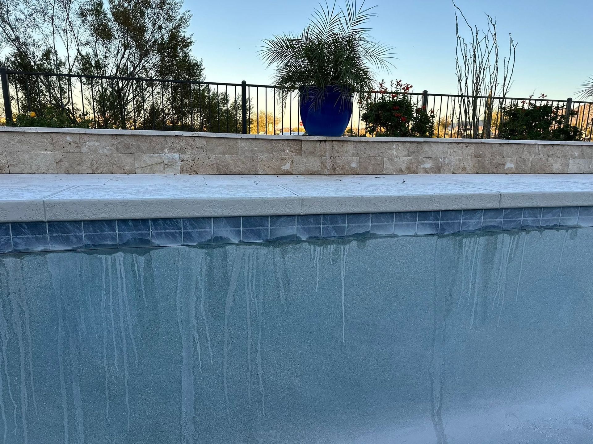Empty blue swimming pool beside a stone deck, with potted plants and trees in the background at sunset