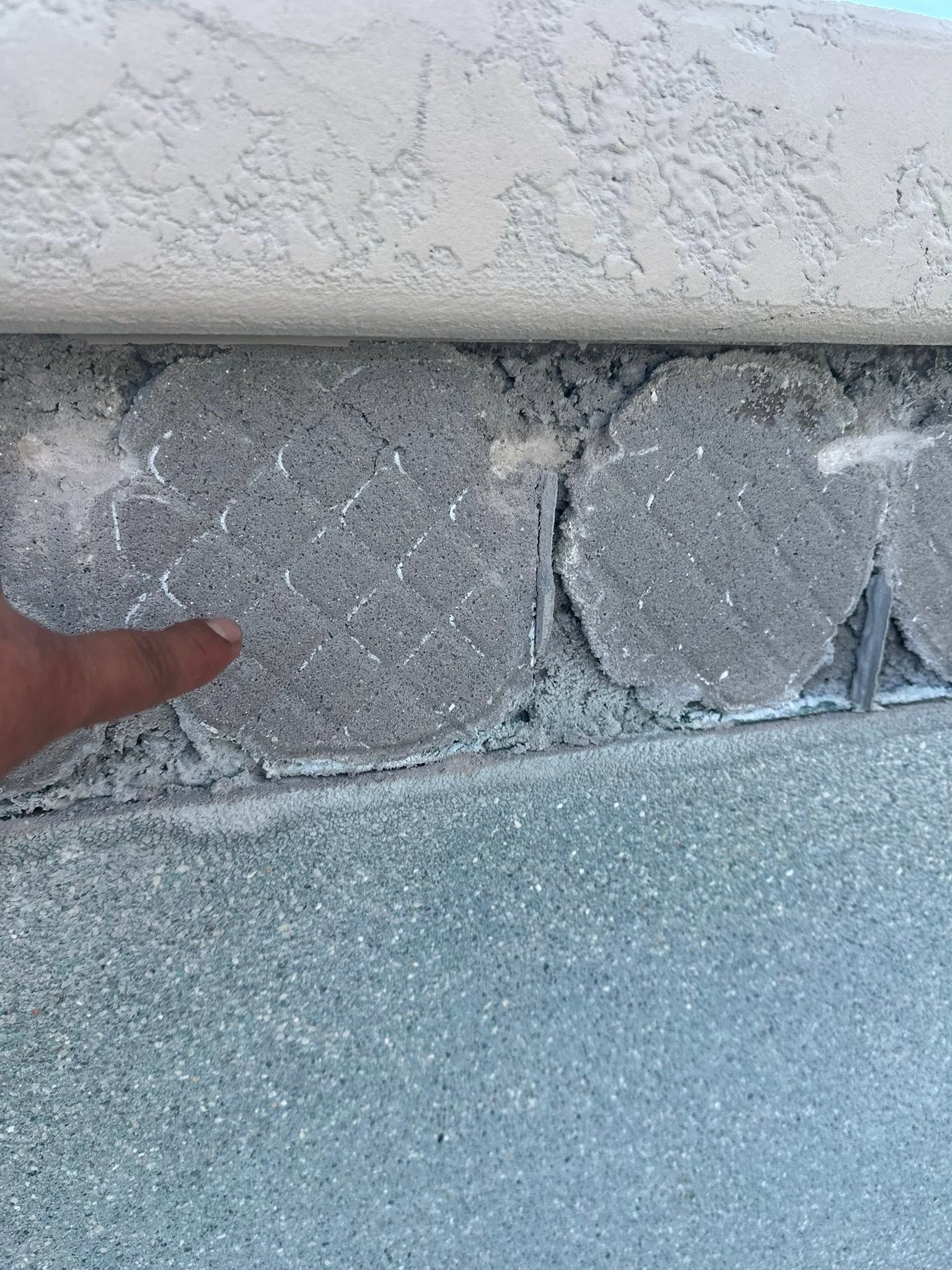Finger pointing at cracked concrete between stucco wall and blue pavement.
