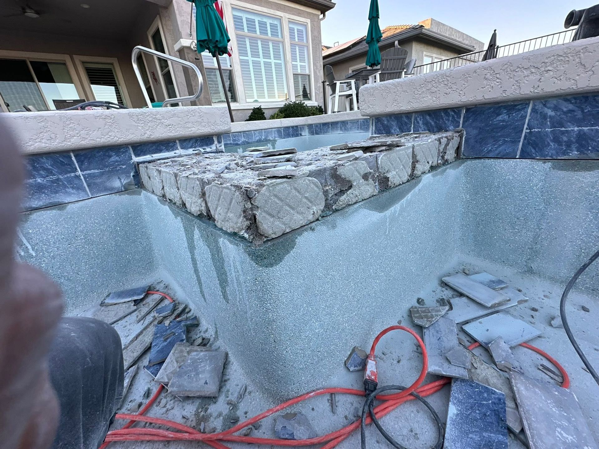 Empty backyard pool with a raised stone platform, exposed wiring, and a house in the background