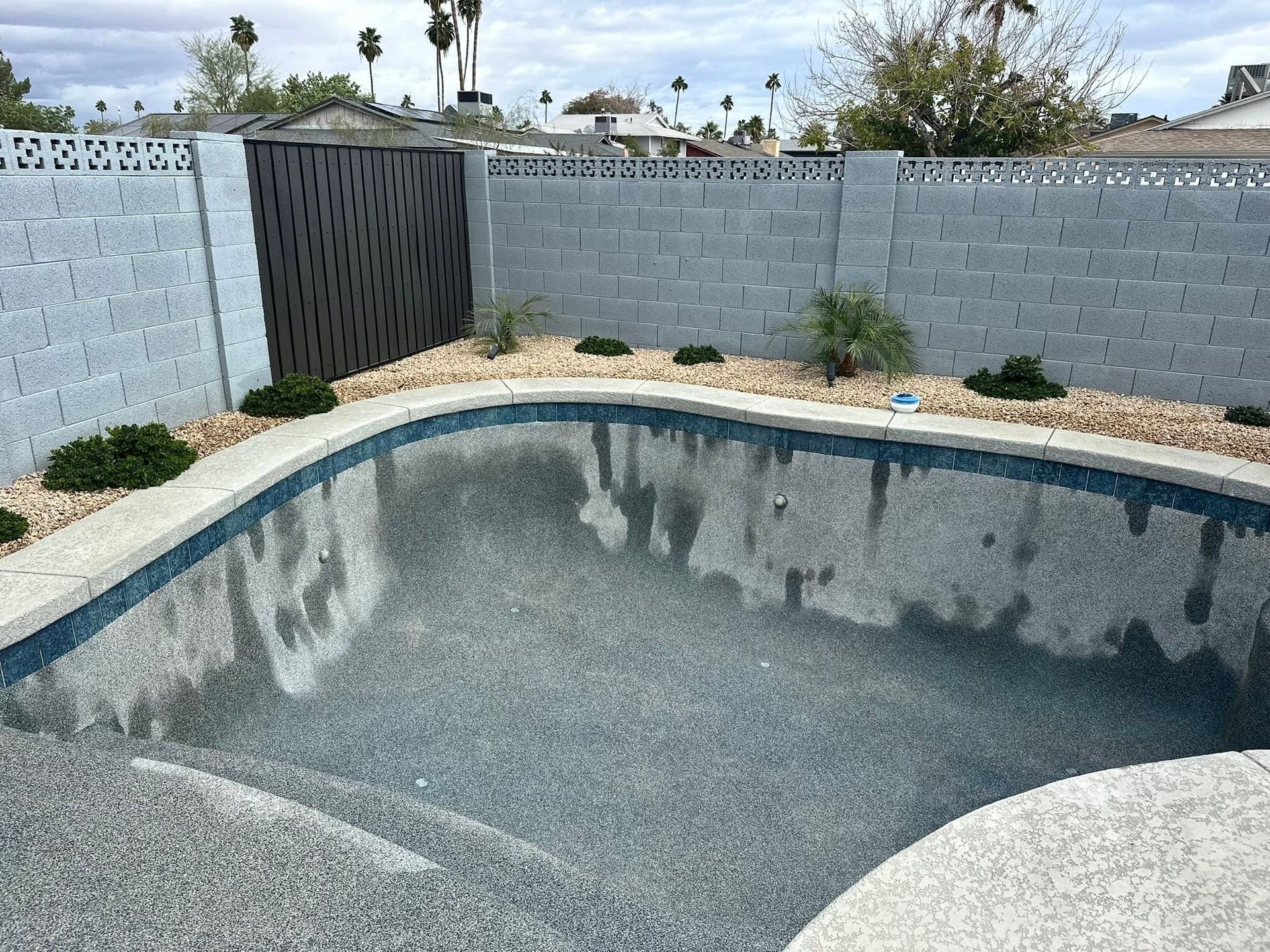 Empty backyard swimming pool with gray walls, surrounded by a concrete fence and desert landscaping