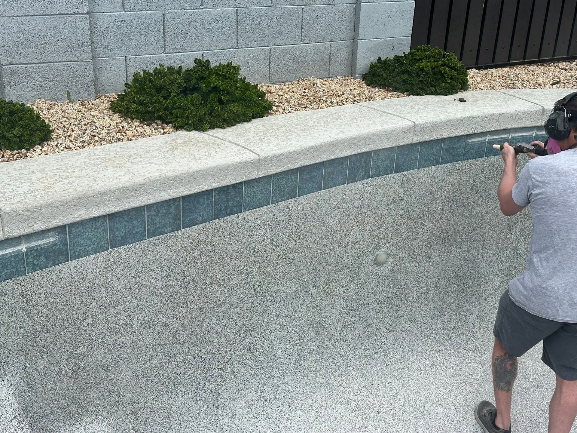 Person using a camera beside an empty pool with a gray wall and shrubs.