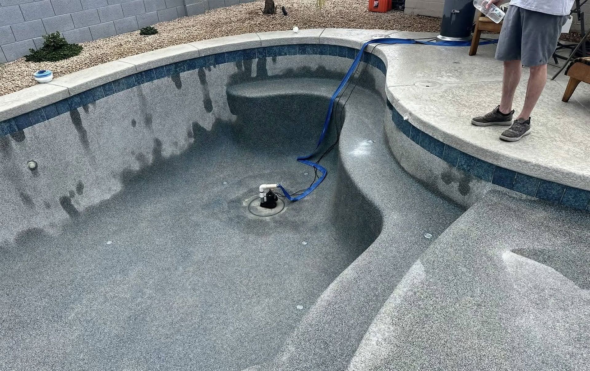 Empty in-ground pool being pressure-washed, with a worker standing by the edge.