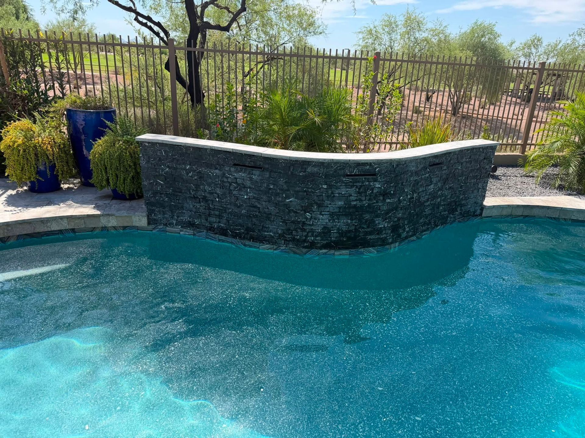 Backyard pool with curved stone water feature and clear blue water, bordered by desert landscaping.