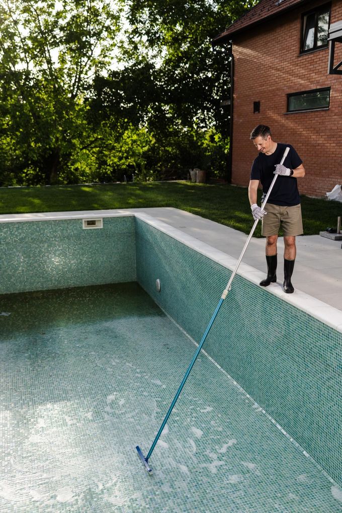 Person using a long pole to clean a tiled swimming pool beside a brick house.
