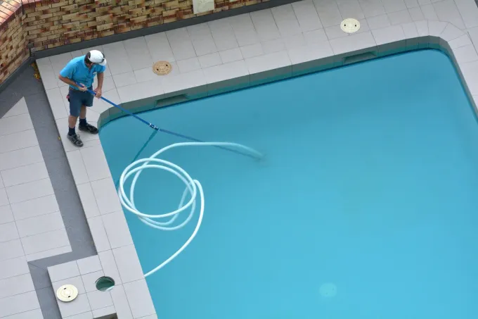 Worker cleaning a swimming pool with a hose, leaving white cleaning lines in the blue water.