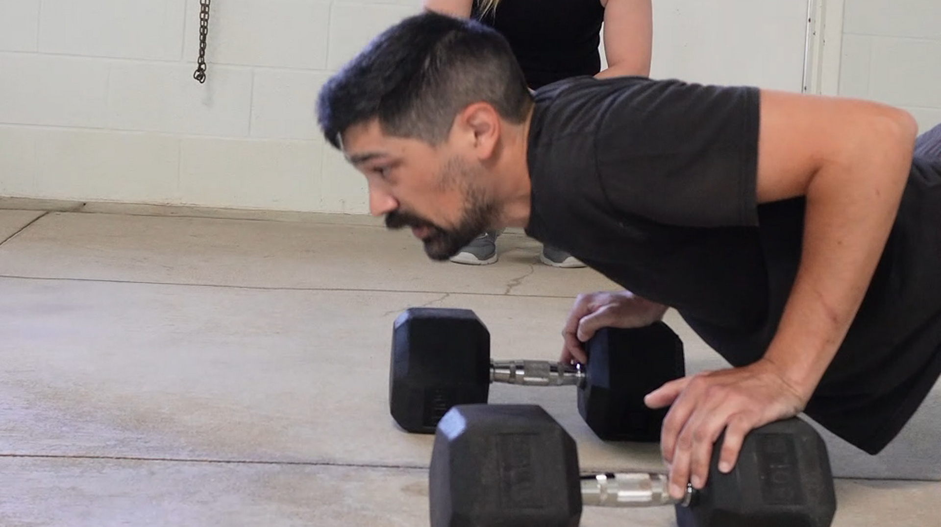 Man doing push-ups on dumbbells in a garage. Another person stands in the background.