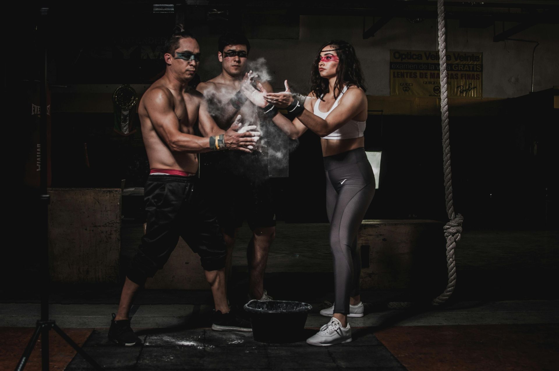 Three people pose with a cloud of white smoke. Dark setting, one shirtless, one in shades, and one in athletic wear.