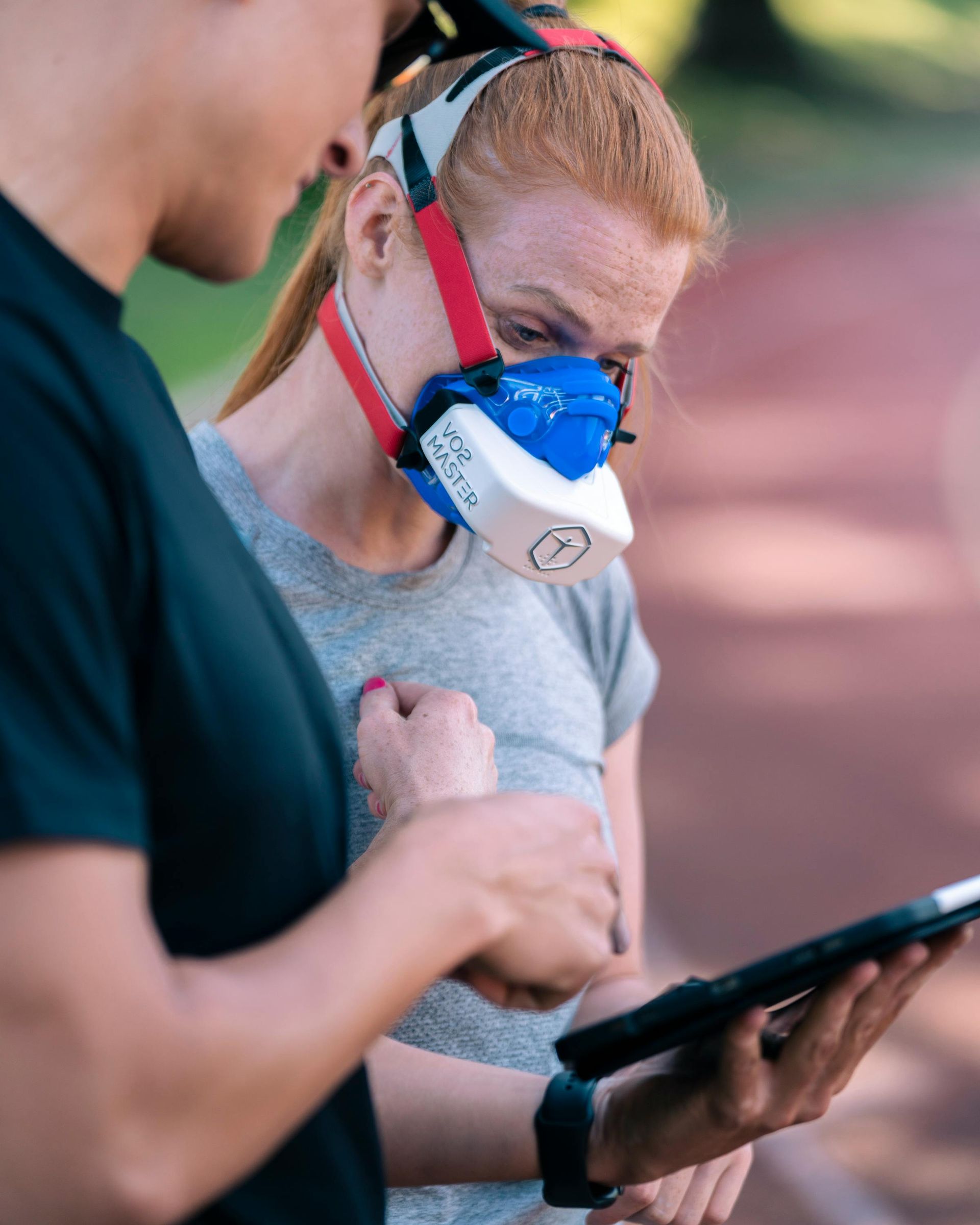 Woman wearing breathing apparatus, consulting a tablet with another person outdoors.
