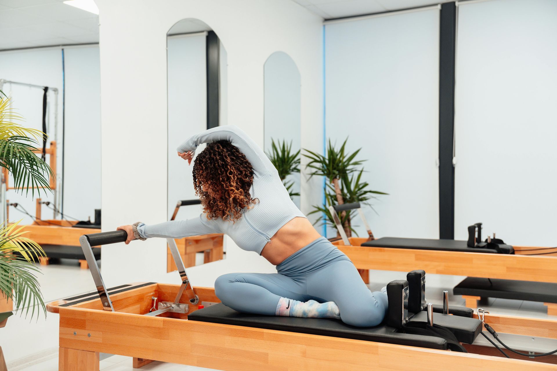 Woman doing Pilates on a reformer, stretching her arm overhead in a light-filled studio.