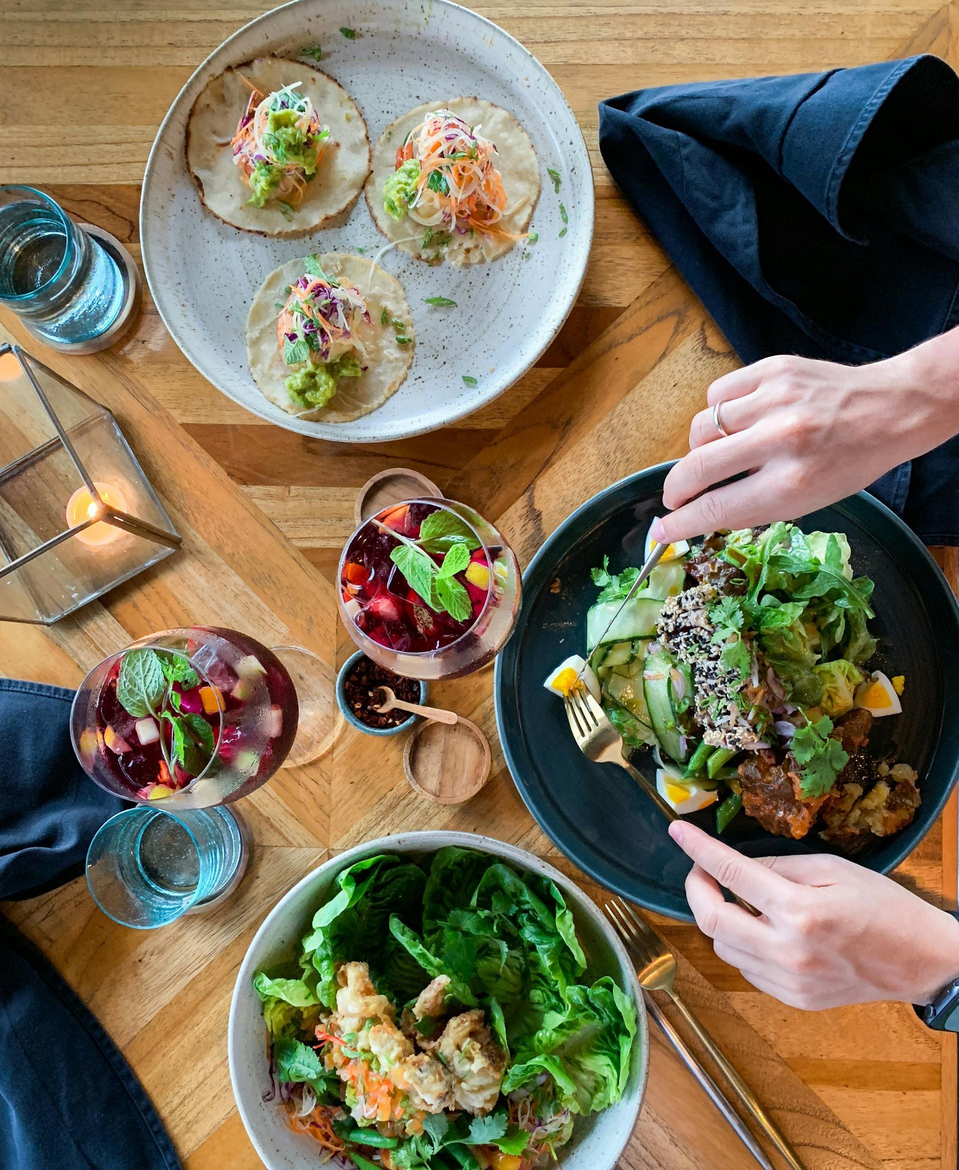 Overhead shot of a table with plates of tacos, salad, and drinks. Hands reach for food.
