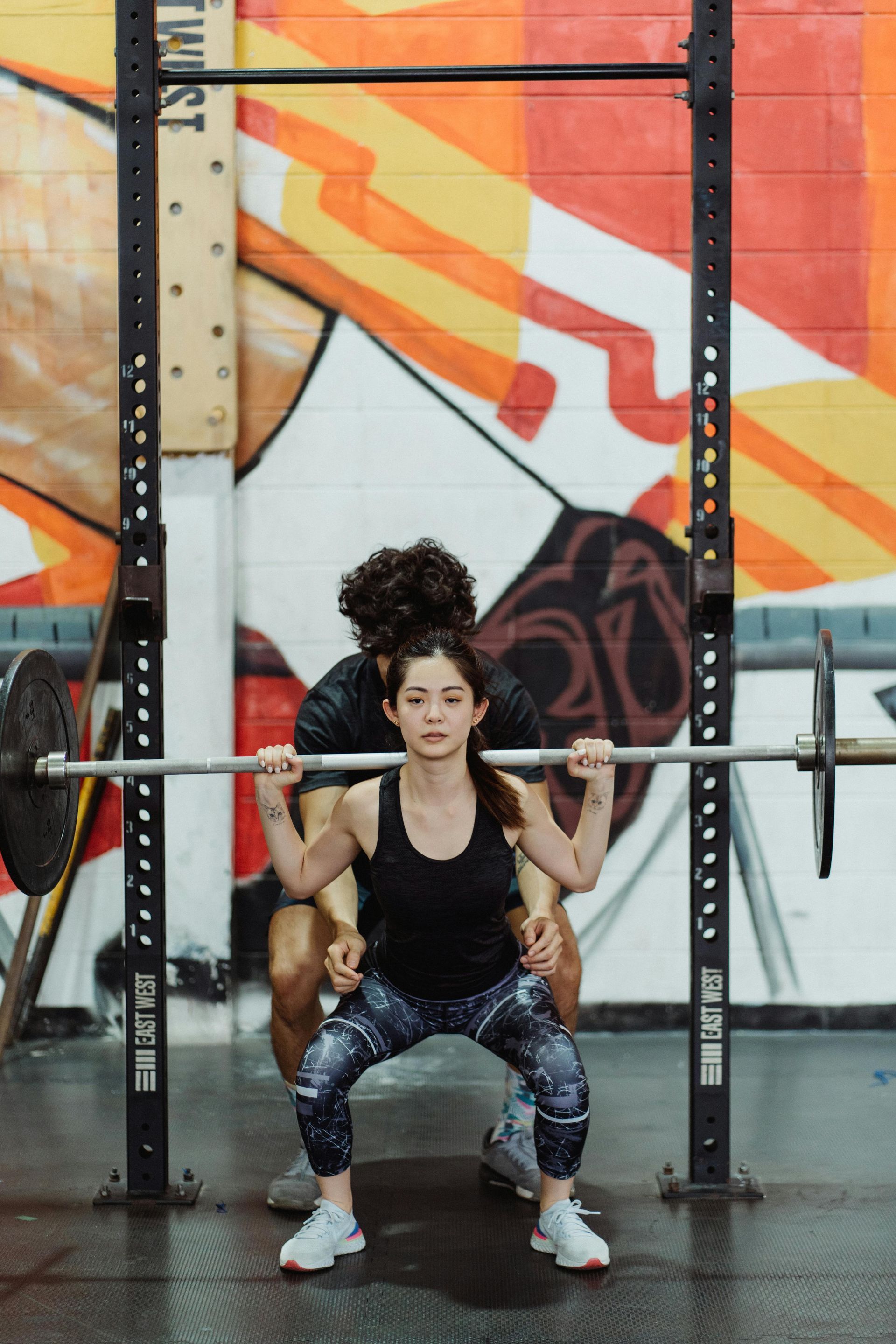 Woman squats with barbell, trainer behind assisting, in a gym with colorful mural on the wall.