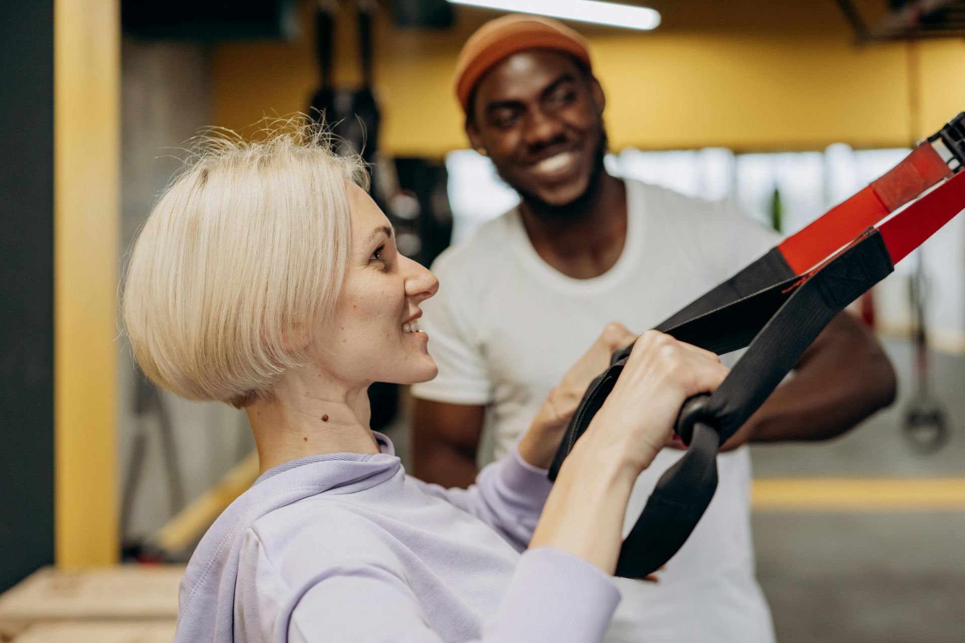 Woman exercising with straps, smiling at a smiling man in a gym. Black and red straps, light clothing.
