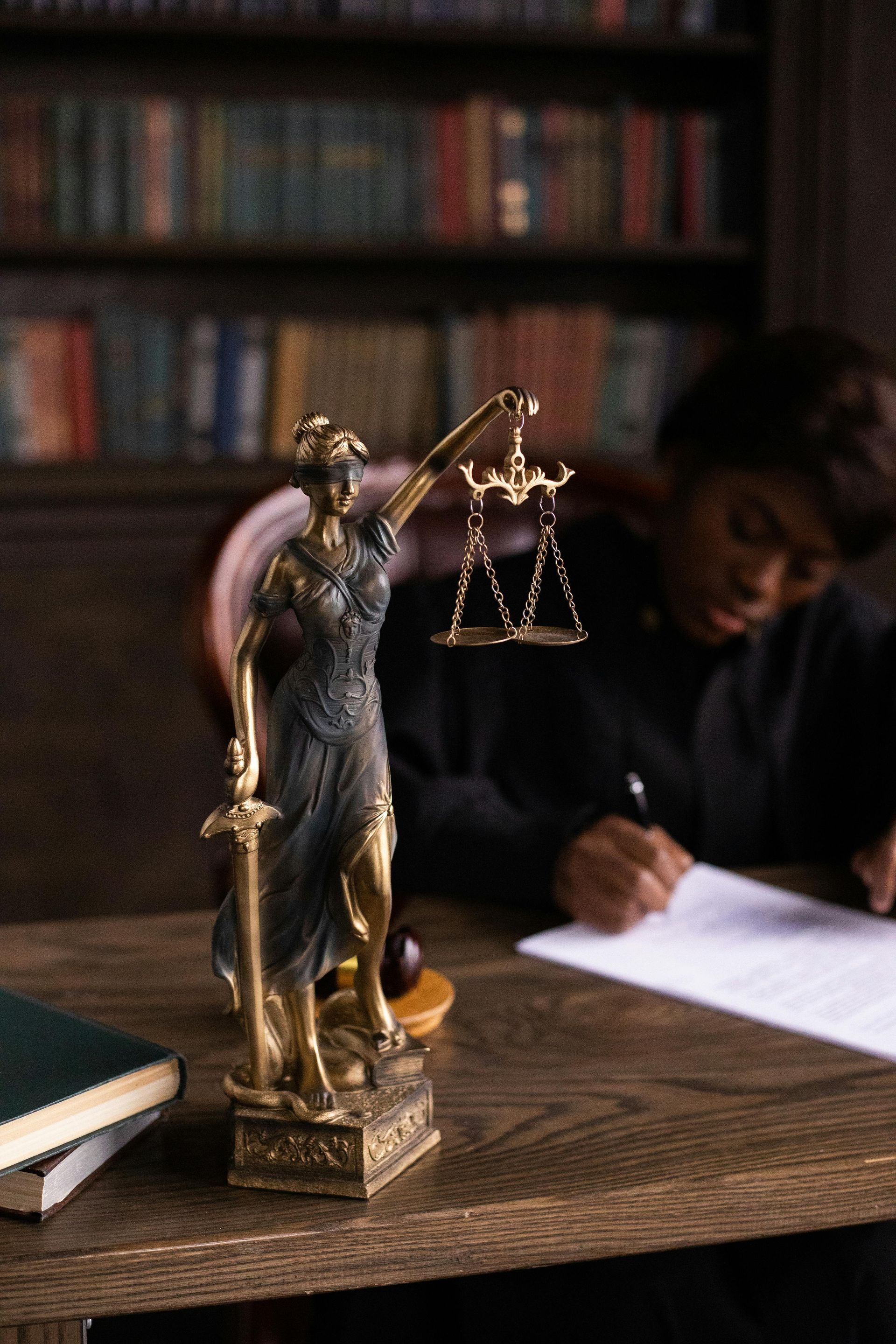 A bronze statue of Lady Justice stands on a desk in front of a person writing in a law library.