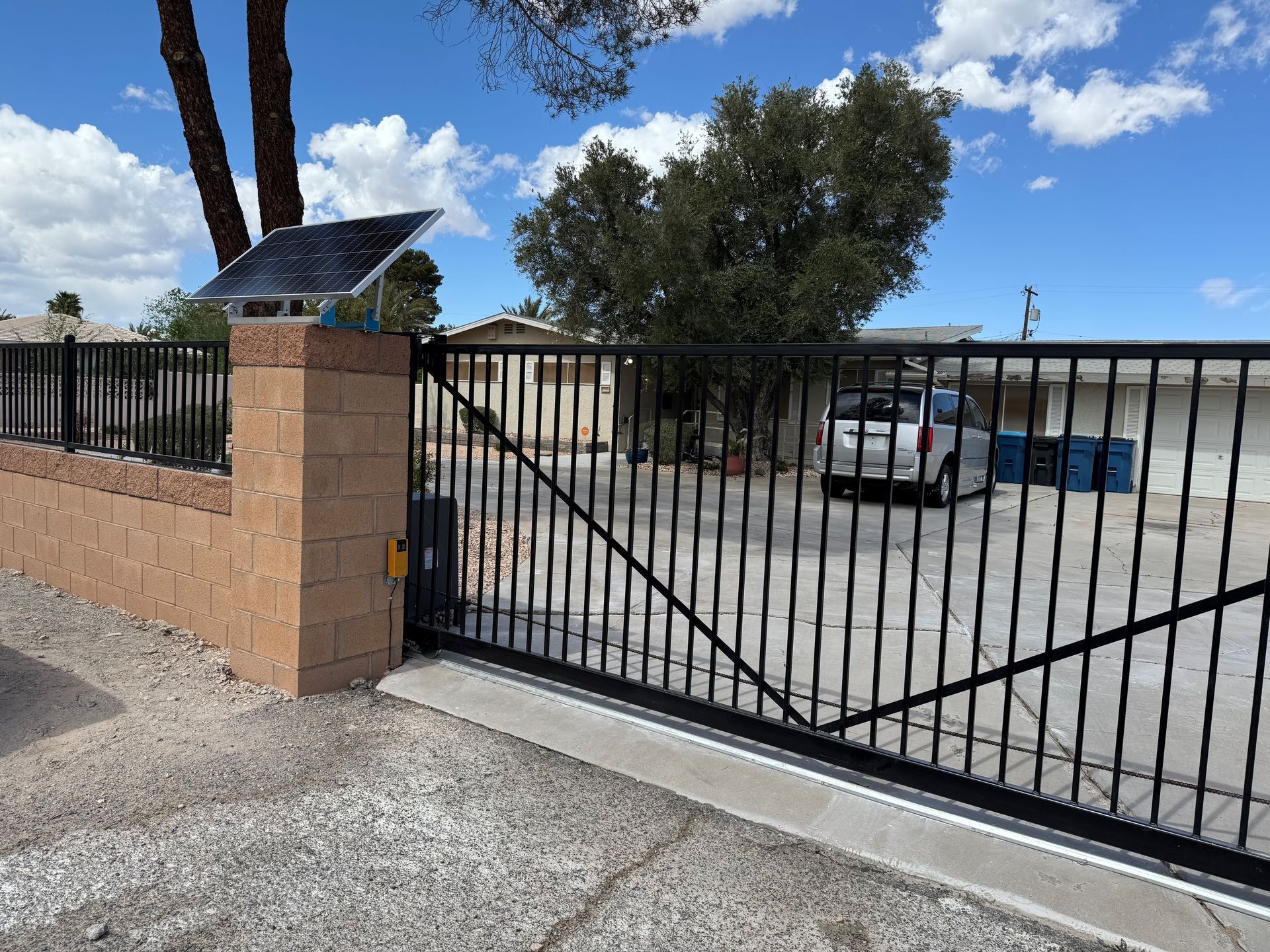 Black iron gates open to a driveway, framed by stone pillars and a house with flower boxes.