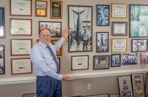 Man standing in an office, surrounded by framed photos, certificates, and aviation memorabilia.