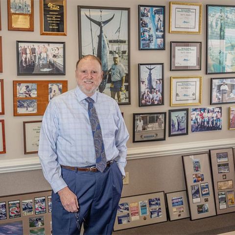 Man standing in an office, surrounded by framed photos, certificates, and aviation memorabilia.
