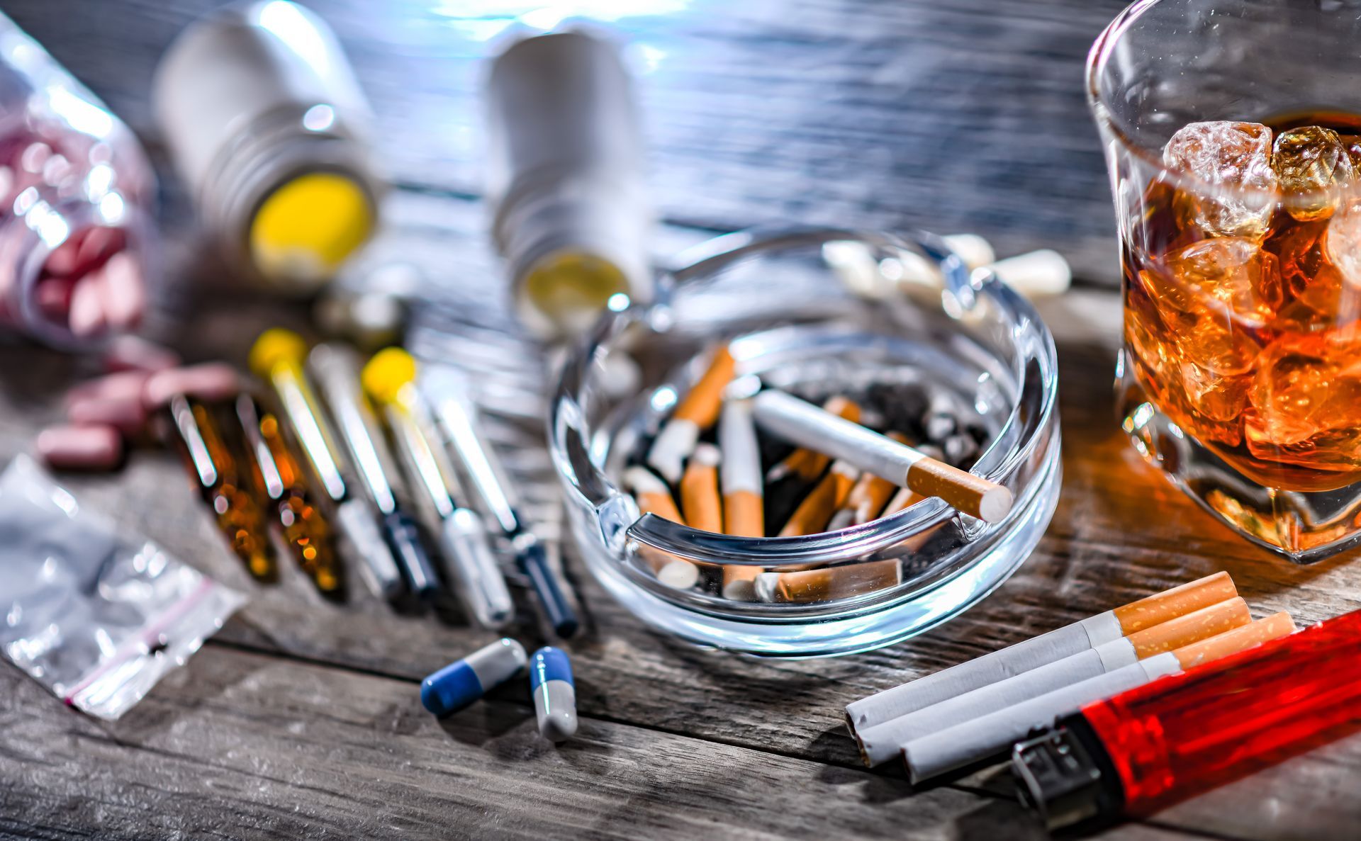Alcohol bottles, scattered pills, syringes, and a glass of whiskey on a wooden table
