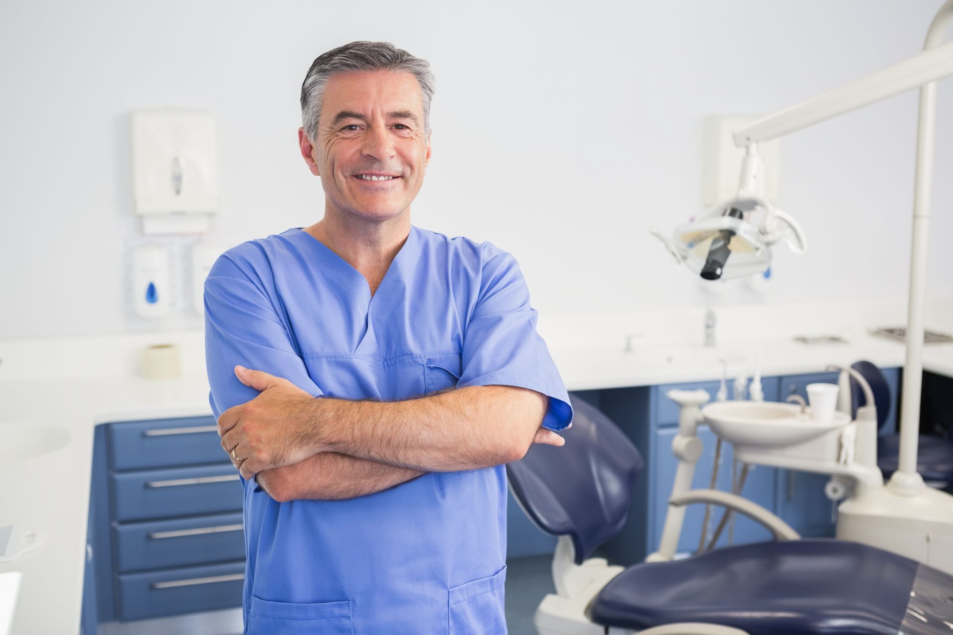 Smiling dentist in blue scrubs standing with arms crossed in a bright treatment room with a chair.