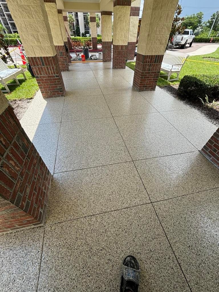 Covered walkway with terrazzo-like tiled floor, brick and beige columns, grass on either side.