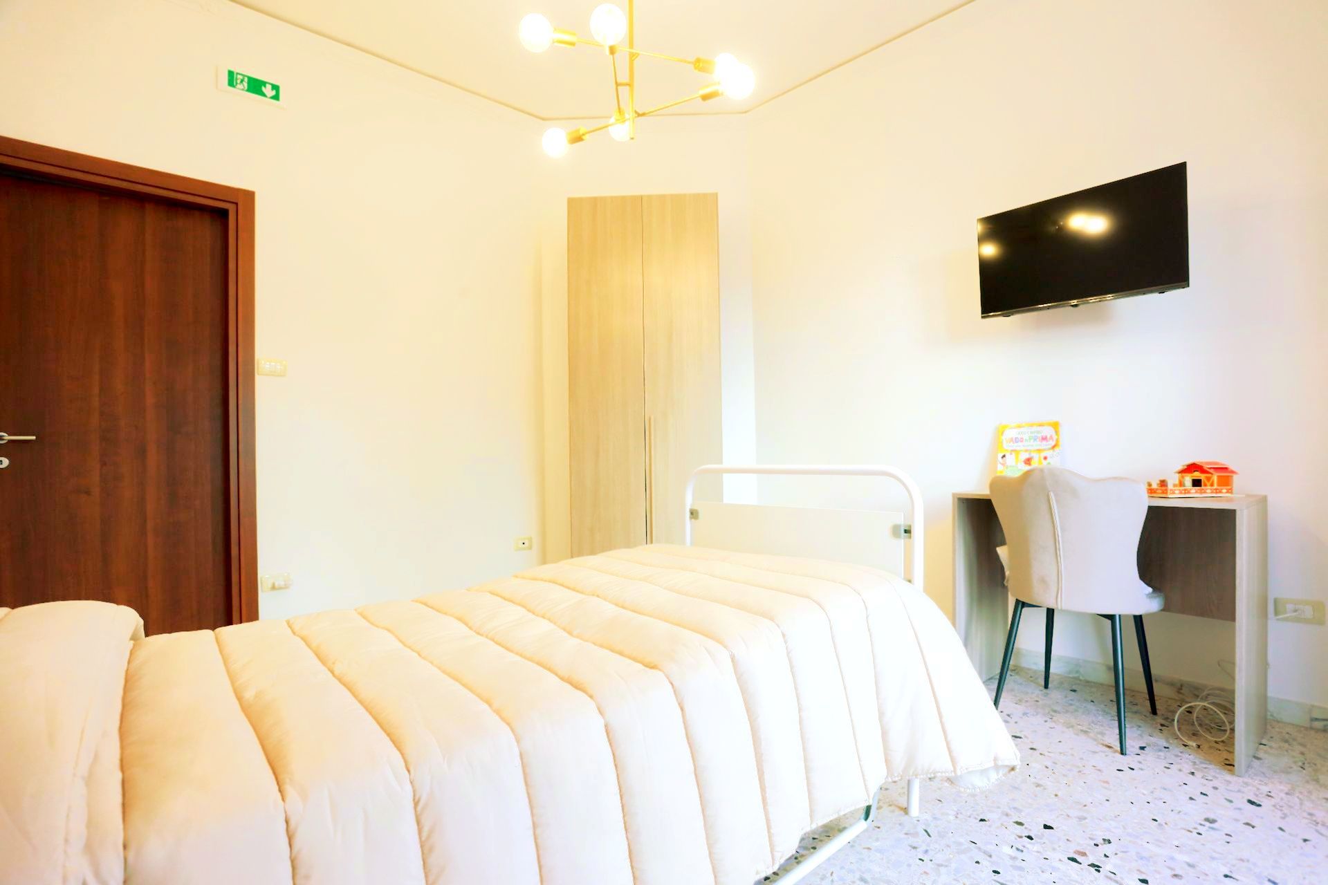 Camera da letto con letto, scrivania, TV e armadio; pareti bianche, porta marrone e lampadario dorato.