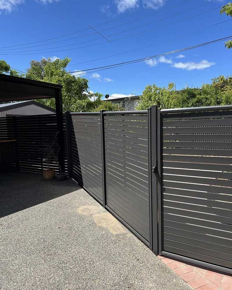 Gray Slat Fence and Gate Enclosing a Paved Driveway With a Dark Carport — CLF Landscapes in Woden, ACT