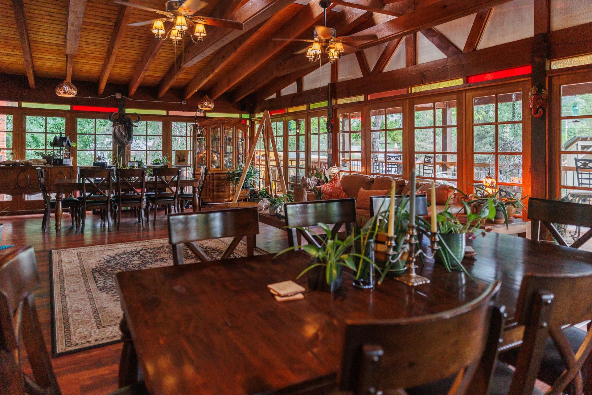 Warm wood-paneled restaurant dining room with tables, chairs, plants, and large windows overlooking greenery
